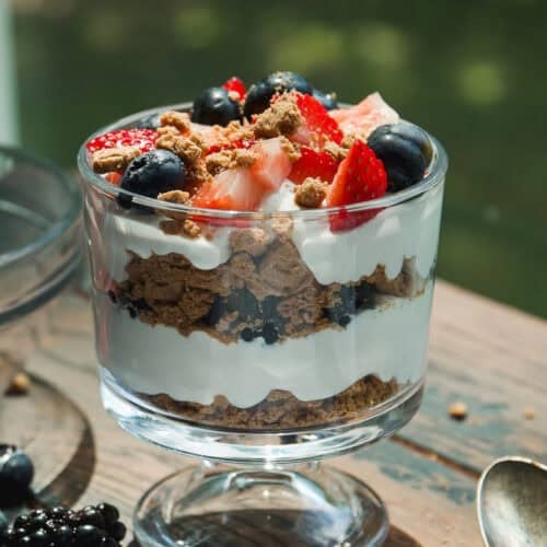 A glass dessert cup filled with layers of whipped cream, crumbled cookies, blueberries, and sliced strawberries, sitting on a wooden table with a spoon and scattered berries nearby.