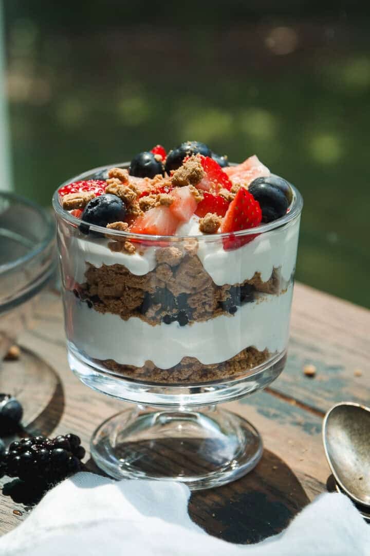 A glass dessert cup filled with layers of whipped cream, crumbled cookies, blueberries, and sliced strawberries, sitting on a wooden table with a spoon and scattered berries nearby.