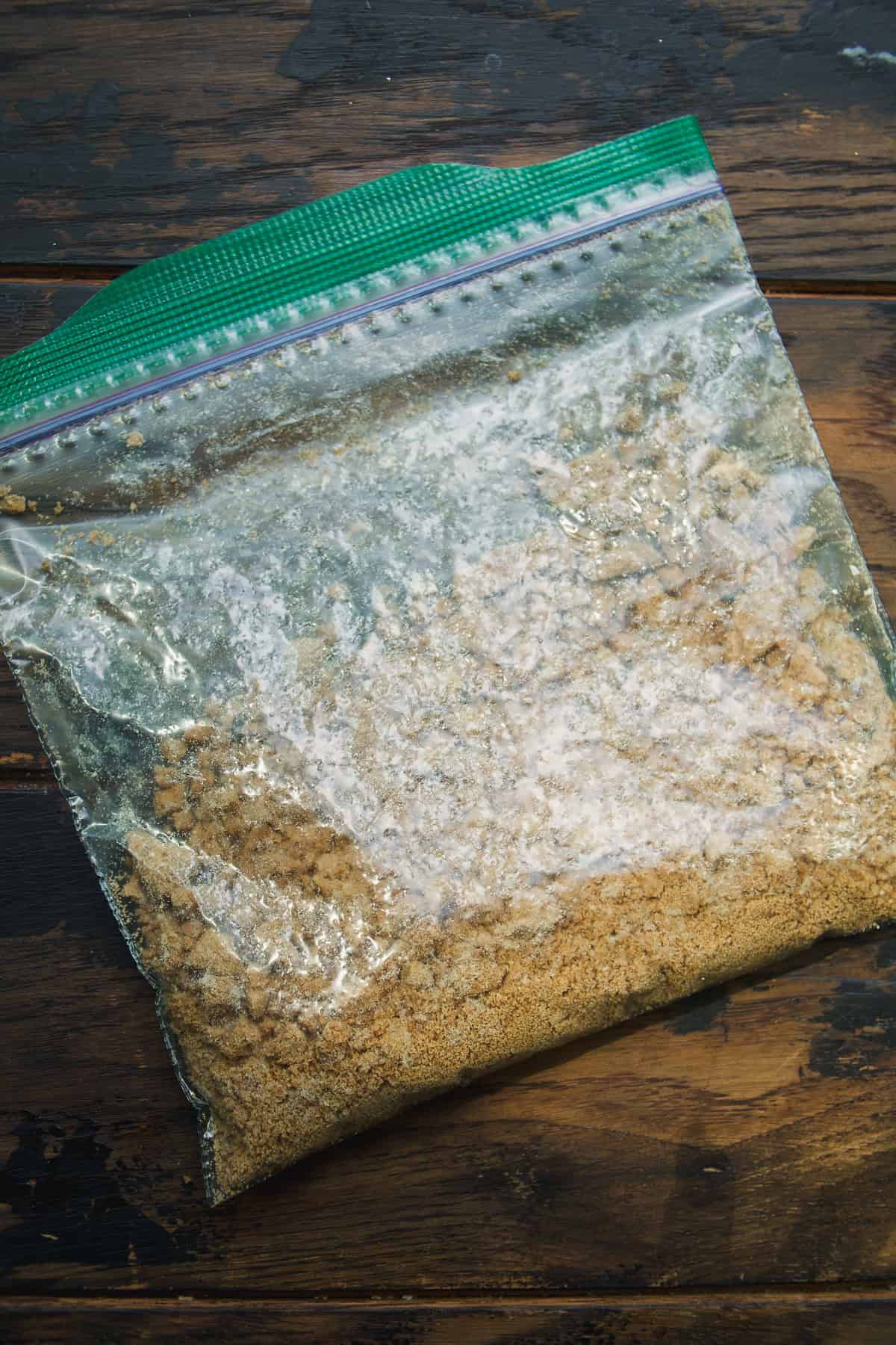 A clear resealable plastic bag filled with crushed graham crackers, resting on a dark wooden surface.