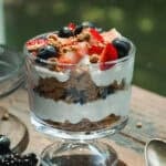 A glass dessert cup filled with layers of yogurt, crumbled cookies, and fresh berries including strawberries and blueberries, sits on a wooden table near a spoon and a white napkin.