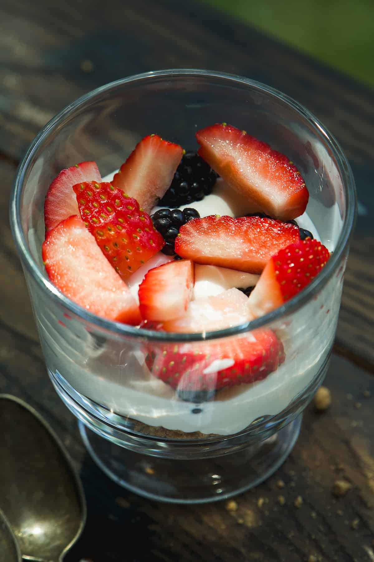 A glass filled with yogurt topped with sliced strawberries and blackberries, placed on a wooden surface with a spoon nearby.