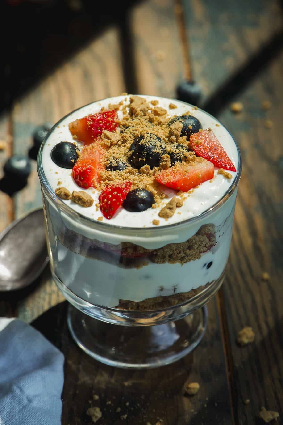 A glass cup filled with layered yogurt, granola, strawberries, and blueberries sits on a wooden table with sunlight streaming in. A spoon and scattered berries are nearby.