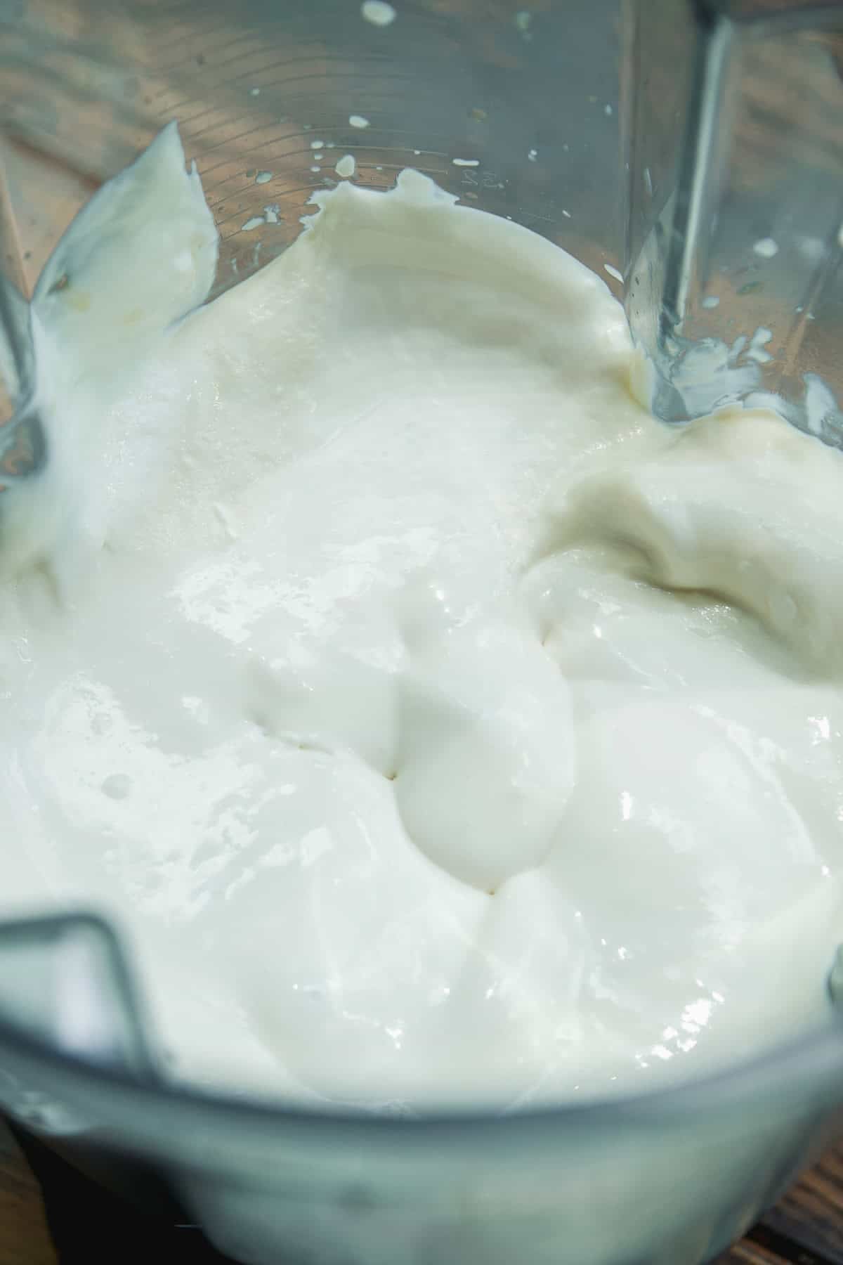 A close-up of thick, creamy white yogurt or a yogurt-like mixture inside a blender container. The texture appears smooth and glossy, with some splatters on the blender sides.