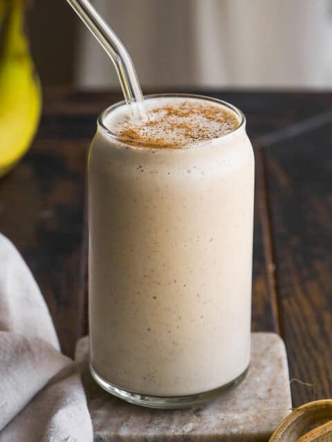 A creamy banana smoothie in a clear glass with a glass straw, topped with a sprinkle of cinnamon, sits on a marble coaster on a rustic wooden table.