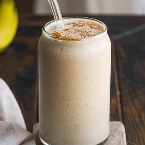 A creamy banana smoothie in a clear glass with a glass straw, topped with a sprinkle of cinnamon, sits on a marble coaster on a rustic wooden table.