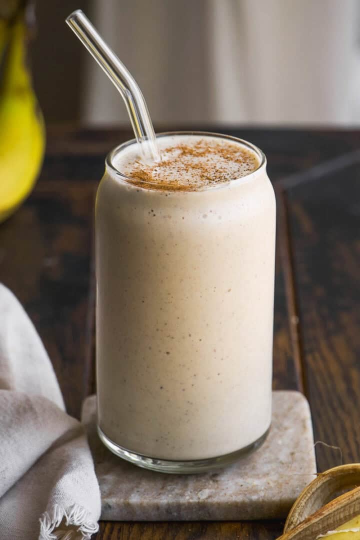 A creamy banana smoothie in a clear glass with a glass straw, topped with a sprinkle of cinnamon, sits on a marble coaster on a rustic wooden table.