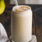 A creamy banana smoothie in a clear glass with a glass straw, topped with a sprinkle of cinnamon, sits on a small marble slab on a wooden table. A beige cloth and a banana are partially visible nearby.