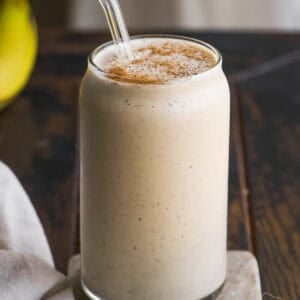 A creamy banana smoothie in a clear glass with a glass straw, topped with a sprinkle of cinnamon, sits on a small marble slab on a wooden table. A beige cloth and a banana are partially visible nearby.
