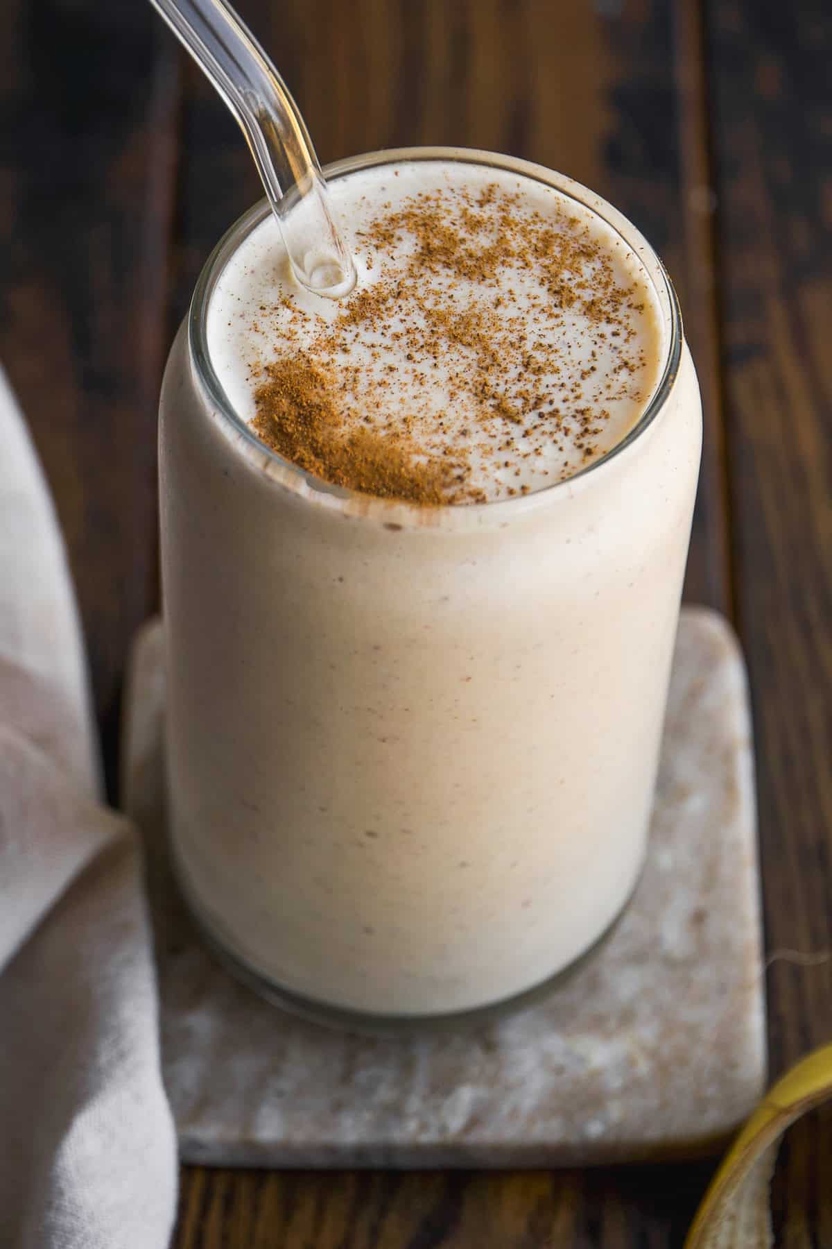 A creamy smoothie topped with a sprinkle of cinnamon sits in a glass with a clear straw, placed on a marble coaster on a wooden table.