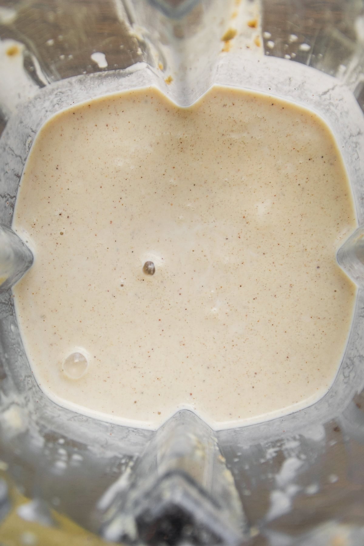 Close-up view of a creamy, pale beige smoothie or batter mixture inside a blender, with small bubbles and specks visible on the surface.
