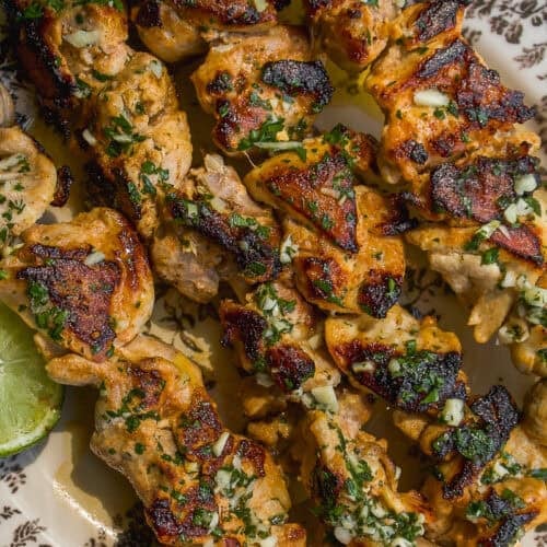 Grilled chicken skewers with a golden-brown char, topped with chopped herbs and garlic, served on a patterned plate with a wedge of lime.