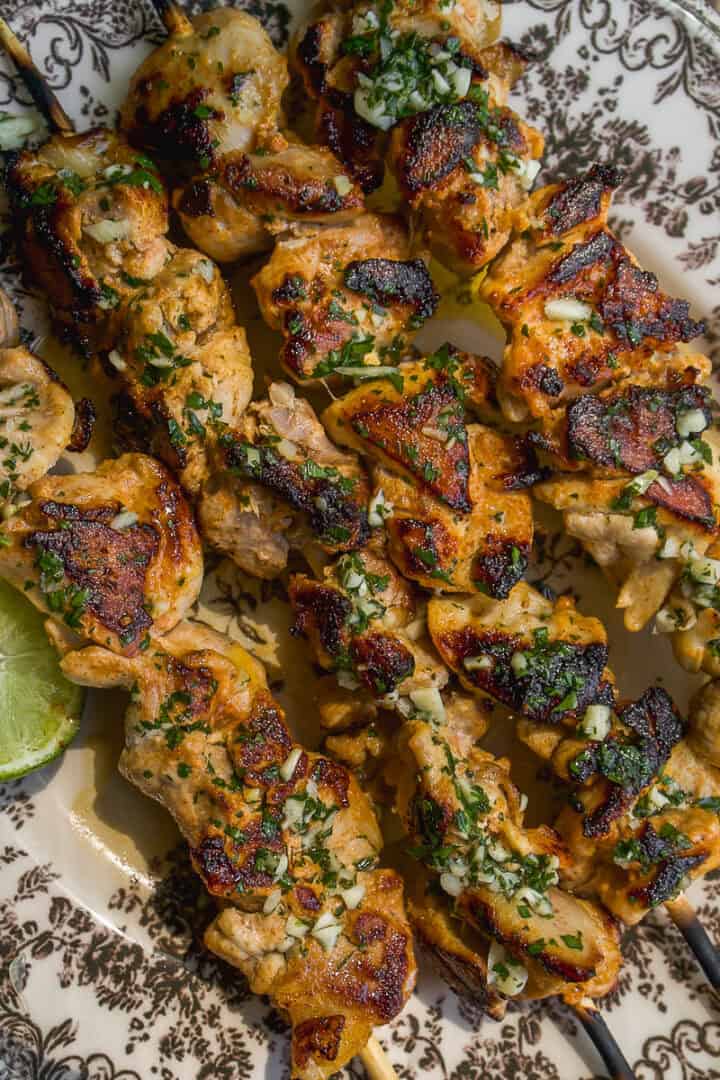 Grilled chicken skewers with a golden-brown char, topped with chopped herbs and garlic, served on a patterned plate with a wedge of lime.