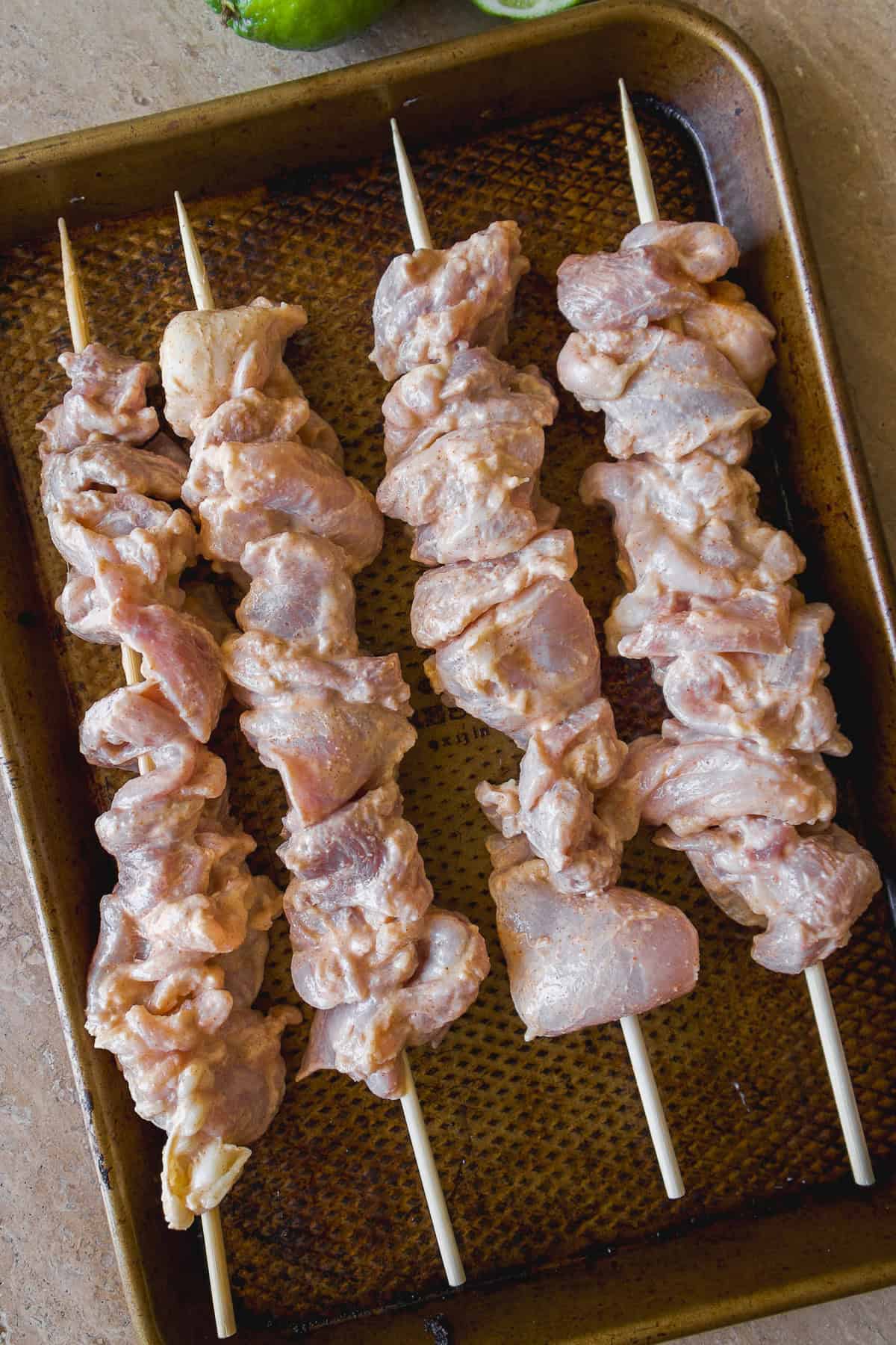 Raw pieces of marinated chicken threaded onto four wooden skewers, arranged on a brown baking sheet, ready for grilling or baking.