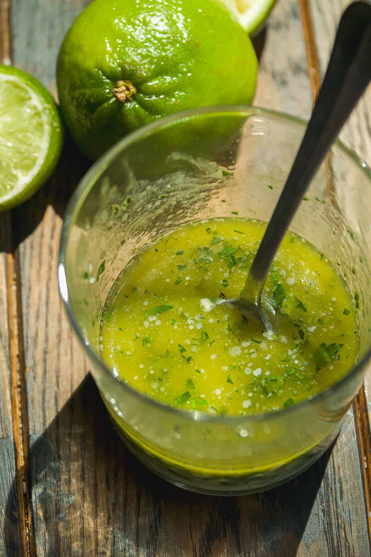 A glass with a spoon containing a green, herbed sauce or dressing sits on a wooden surface, next to a whole lime and a halved lime.