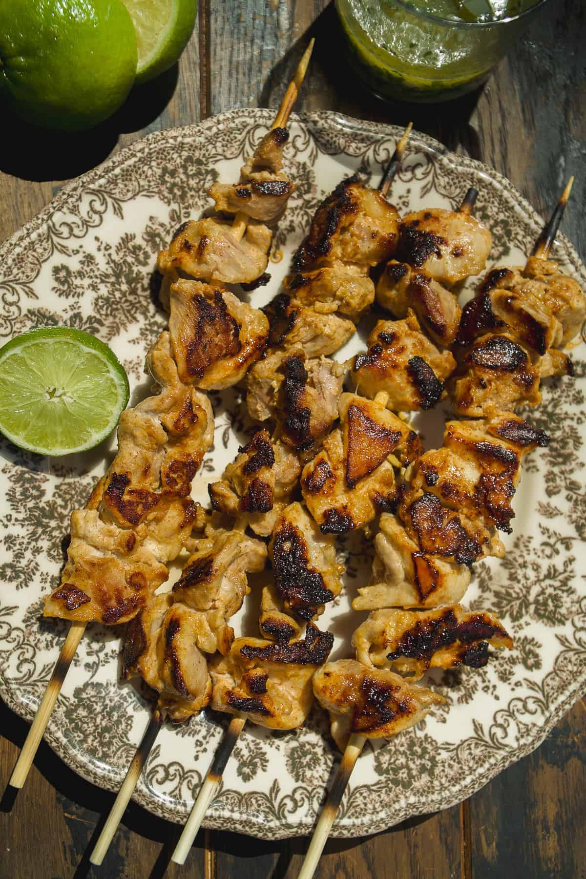 Four grilled chicken skewers are arranged on a patterned plate with a halved lime on the side. Fresh limes and a green beverage are nearby on a rustic wooden table.