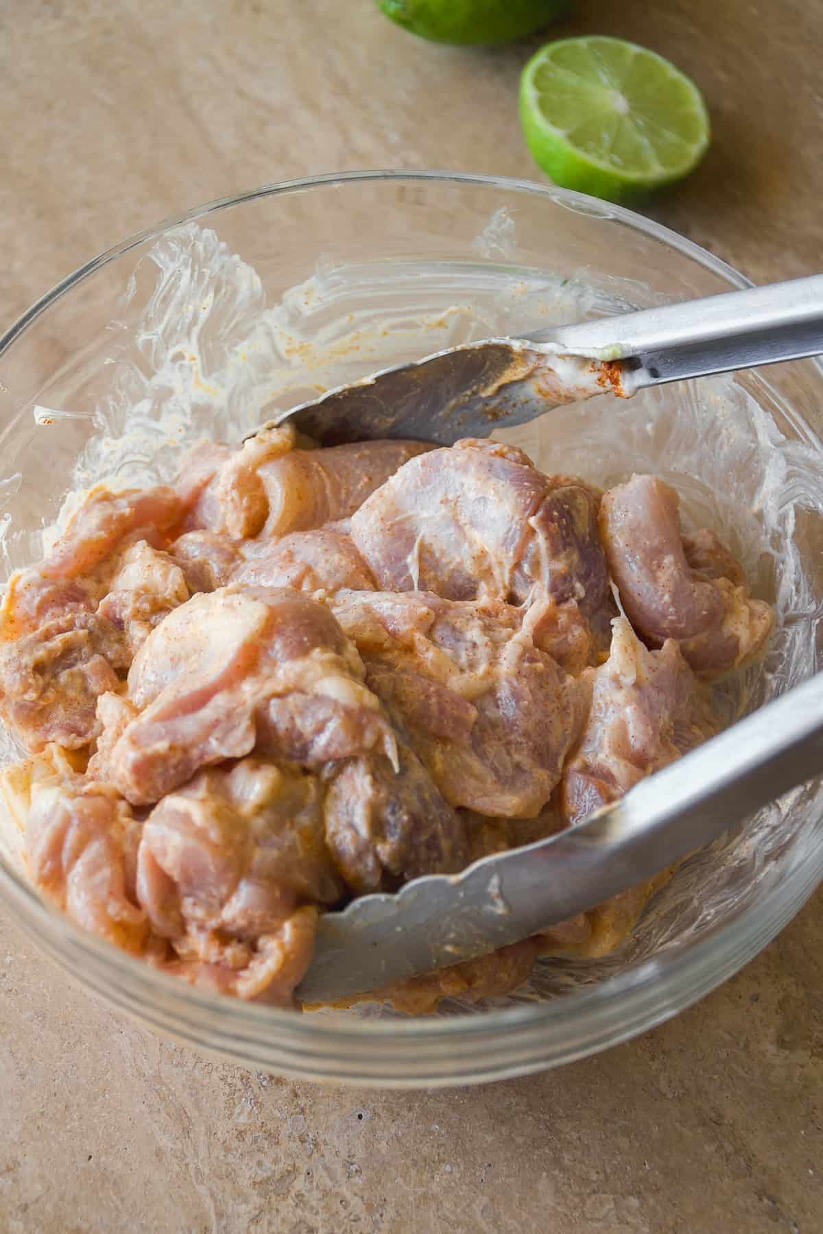 A glass bowl filled with marinated raw chicken pieces being mixed with metal tongs; a halved lime is visible in the background on a brown surface.