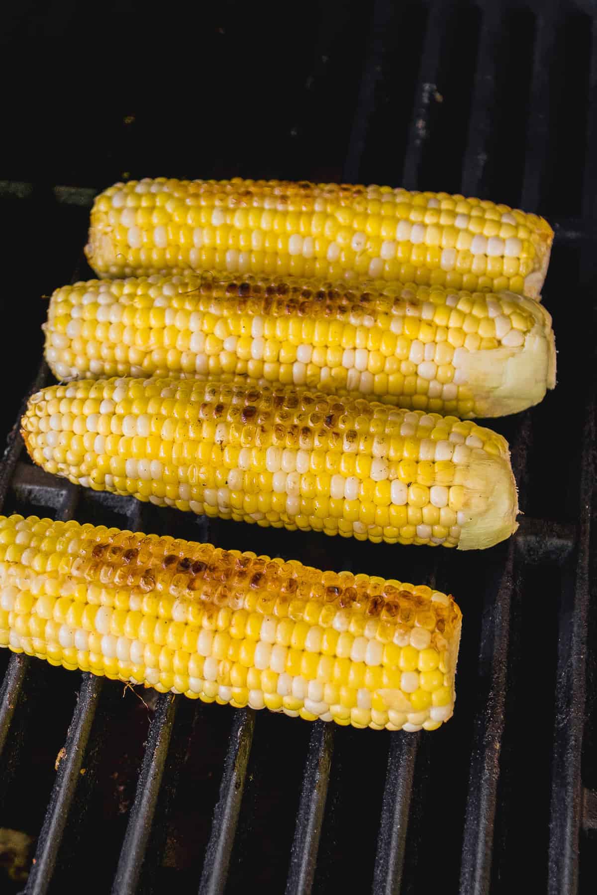 Four ears of corn on the cob are grilling on a barbecue, with visible char marks on the yellow kernels.