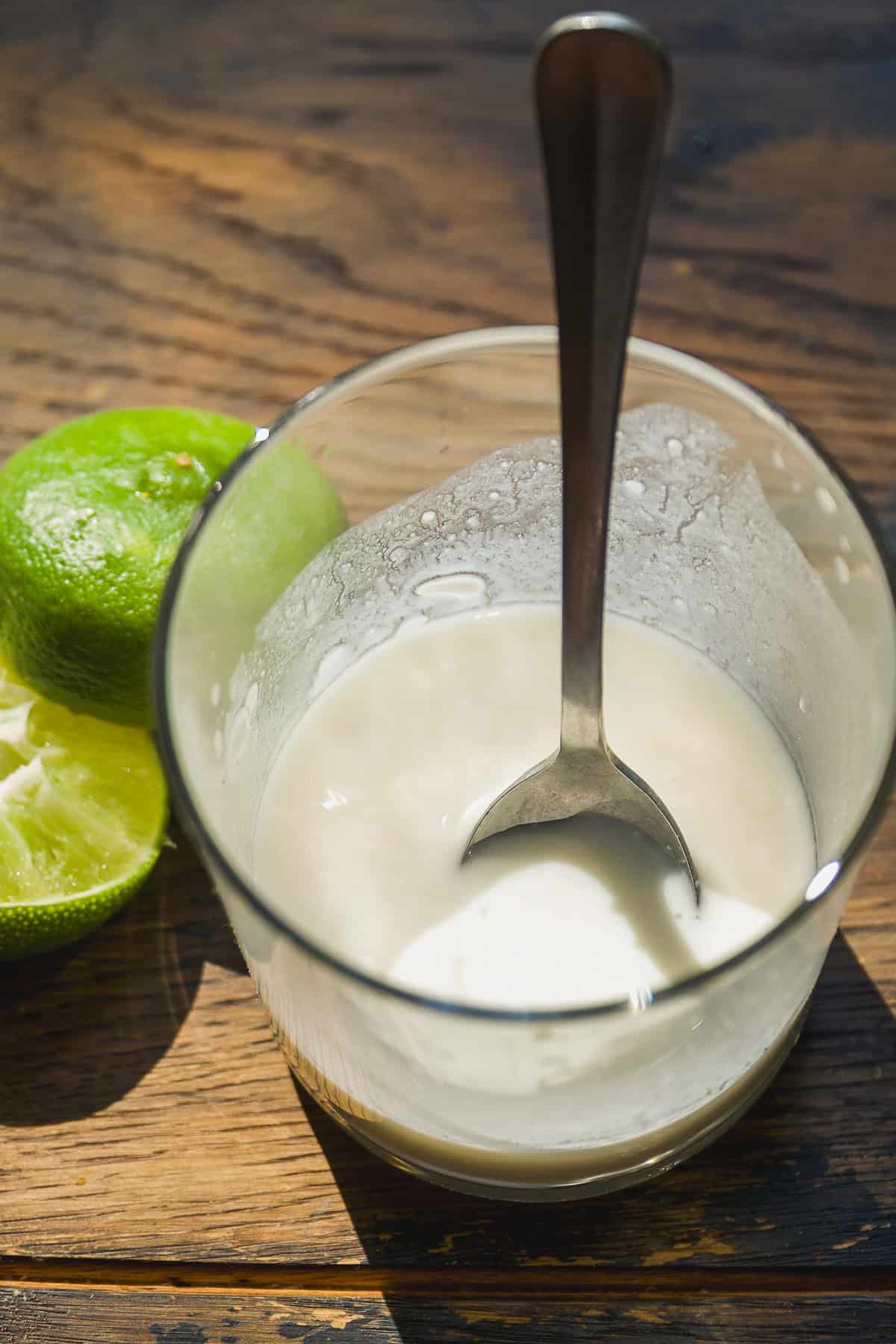 A glass with a creamy white mixture and a spoon inside sits on a wooden surface next to a halved lime.