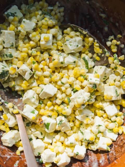 A close-up of a fresh corn salad with diced feta cheese, chopped herbs, and a creamy dressing, all mixed together in a wooden bowl with a metal spoon.