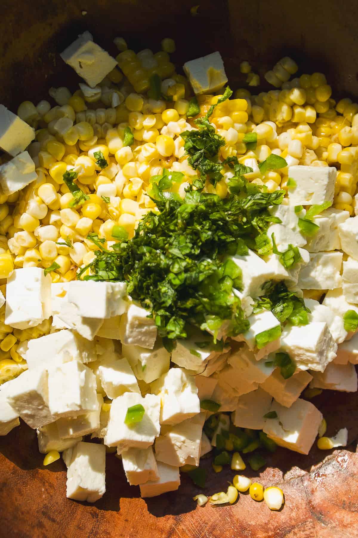 A close-up of a bowl containing diced white cheese, kernels of yellow corn, and chopped green herbs and peppers, all ready to be mixed together.