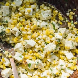 A close-up of a salad featuring yellow corn kernels, cubes of white cheese, fresh herbs, and a creamy dressing, with a metal spoon visible in a wooden bowl.