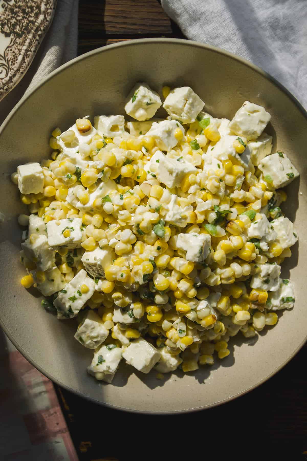 A bowl of corn salad with cubes of white cheese, yellow corn kernels, fresh herbs, and a creamy dressing, served in a shallow beige dish on a wooden table.