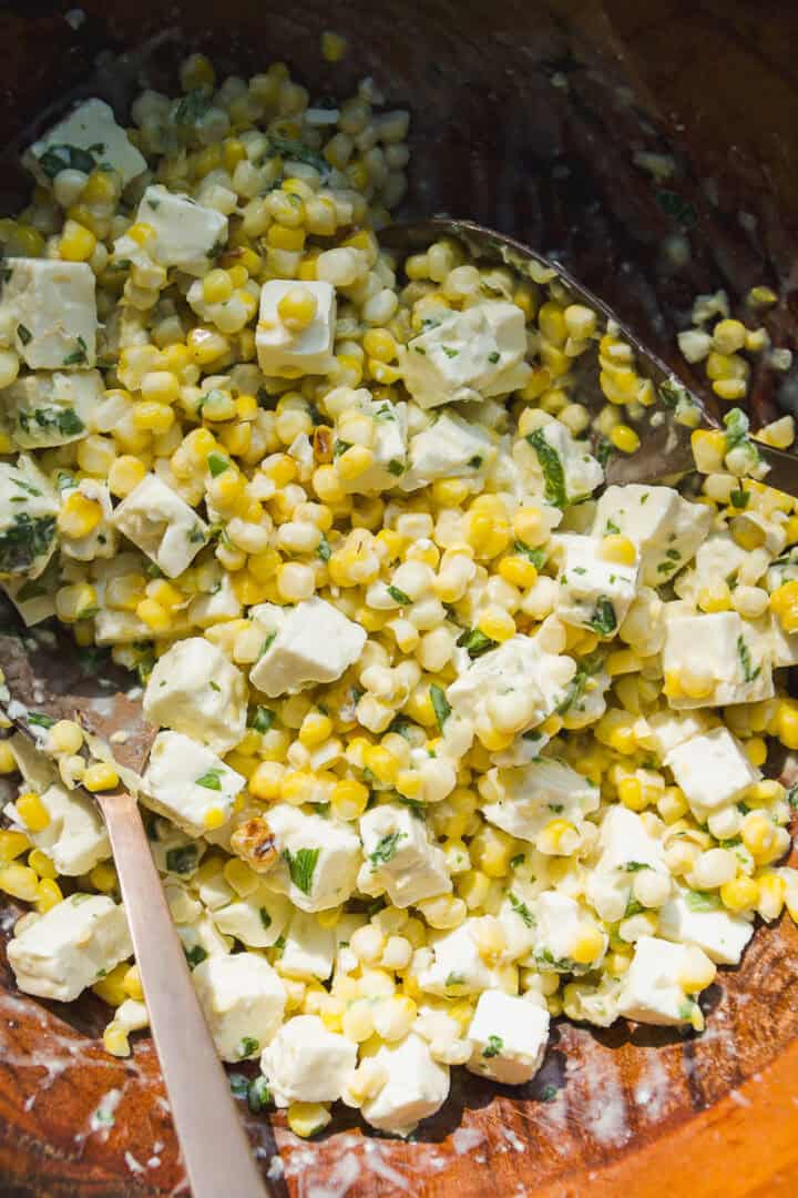 A close-up of a fresh corn salad with diced feta cheese, chopped herbs, and a creamy dressing, all mixed together in a wooden bowl with a metal spoon.