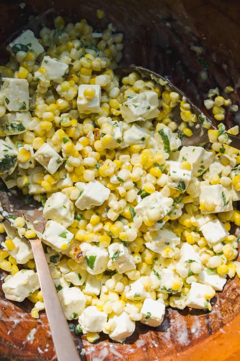 A close-up of a fresh corn salad with diced feta cheese, chopped herbs, and a creamy dressing, all mixed together in a wooden bowl with a metal spoon.