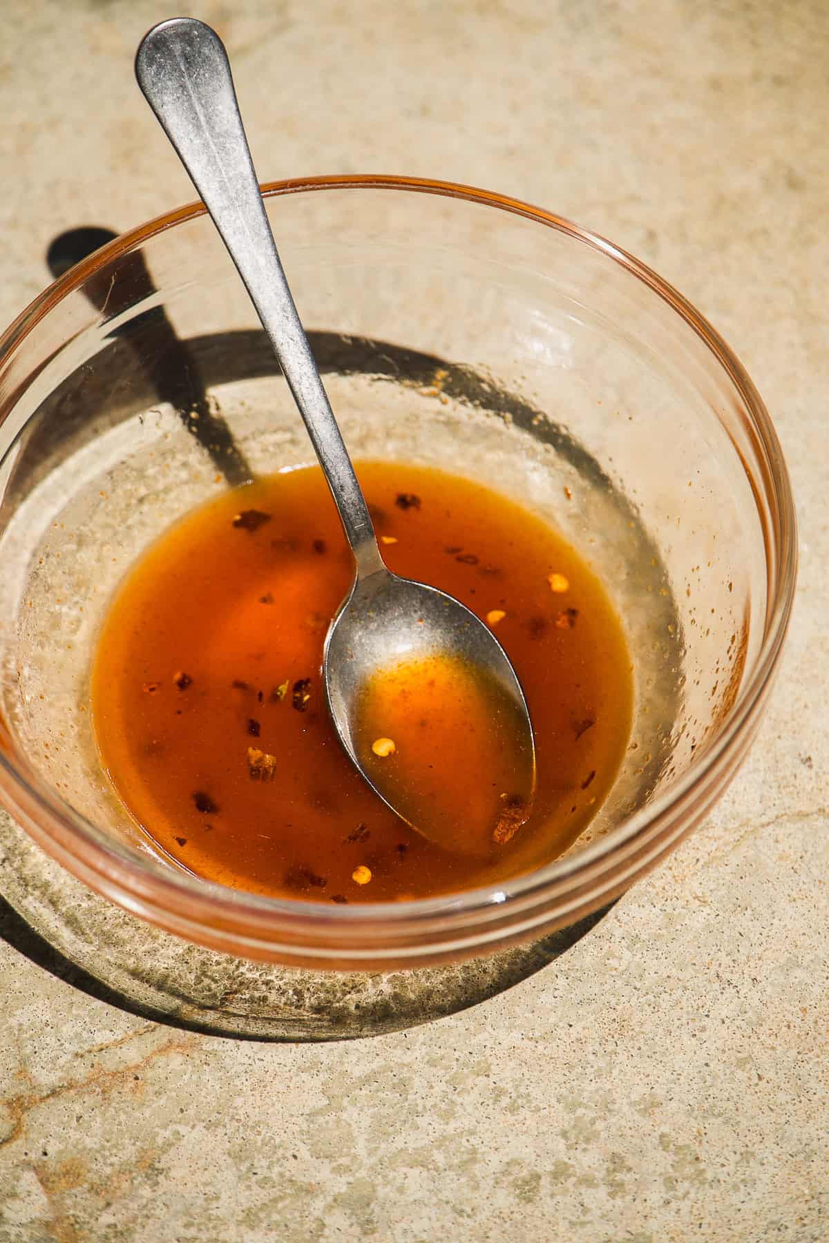 A glass bowl with a metal spoon containing a small amount of reddish-brown liquid with visible chili flakes and bits of seasoning, placed on a light-colored textured surface.