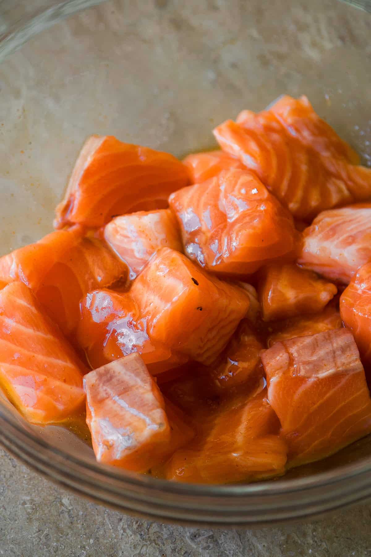 Raw salmon chunks in a glass bowl, marinated and ready for cooking, on a beige countertop.