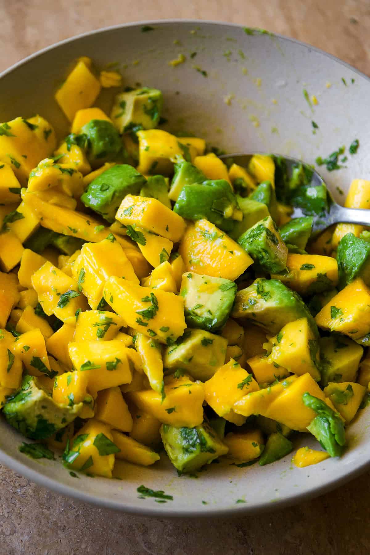 A bowl filled with a colorful mixture of diced mango, avocado, and chopped cilantro, with a spoon resting inside. The ingredients are fresh and vibrant, creating a healthy salad.