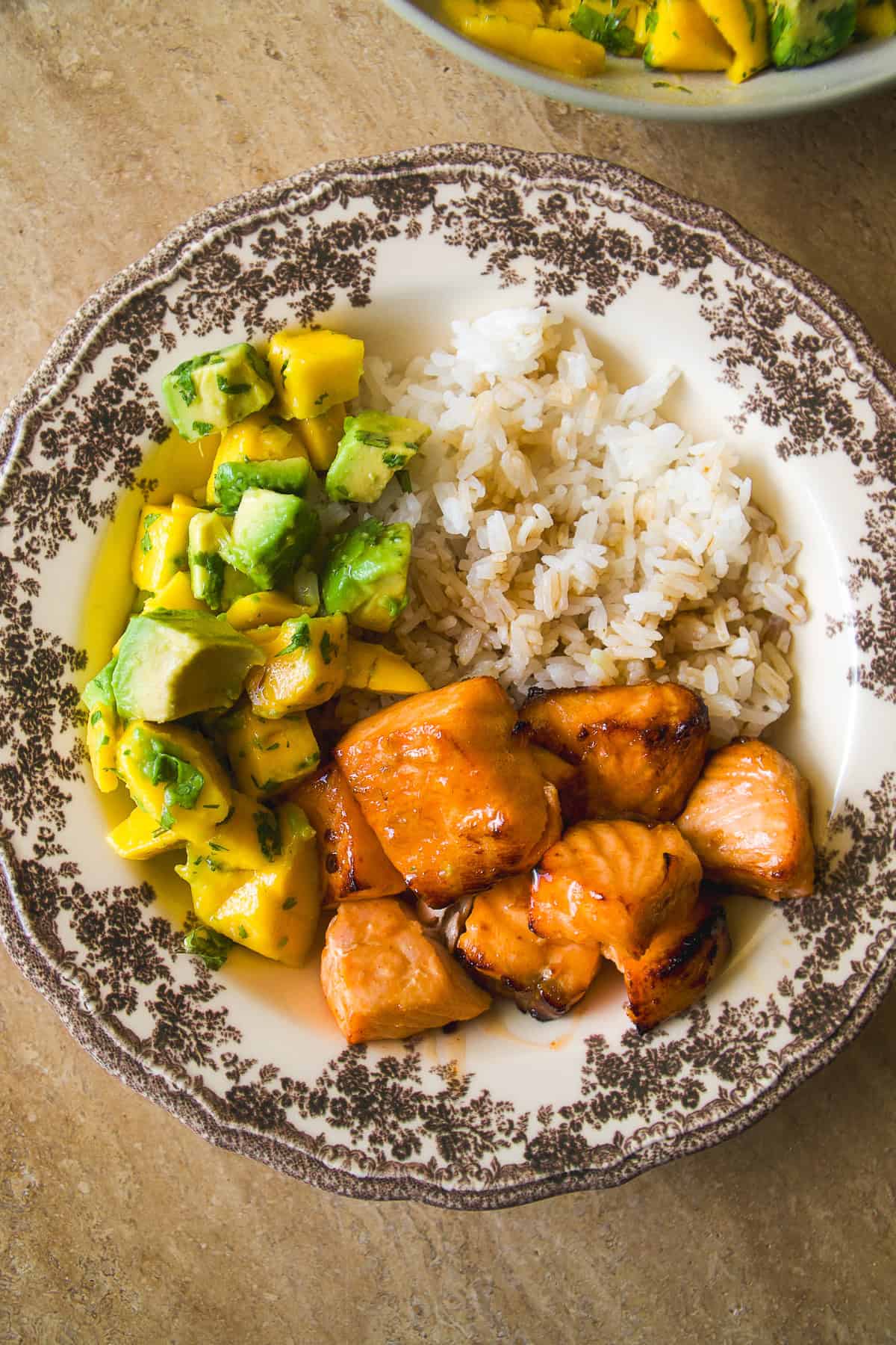 A decorative plate with white rice, glazed salmon pieces, and a mango avocado salsa, arranged in separate sections on a tan surface.