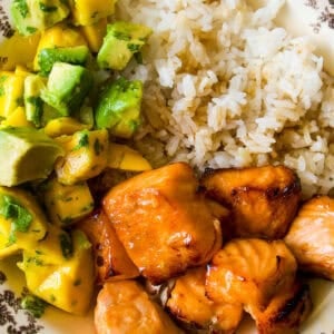 A plate with white rice, grilled salmon pieces, and a mango avocado salsa garnished with herbs, served on a vintage-patterned dish.
