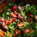 A wooden bowl filled with a fresh salad of leafy greens, sliced strawberries, pecans, red onions, and crumbled cheese, mixed with a wooden spoon.