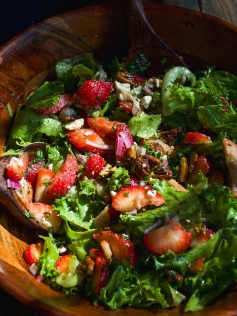 A wooden bowl filled with a fresh salad of leafy greens, sliced strawberries, pecans, red onions, and crumbled cheese, mixed with a wooden spoon.