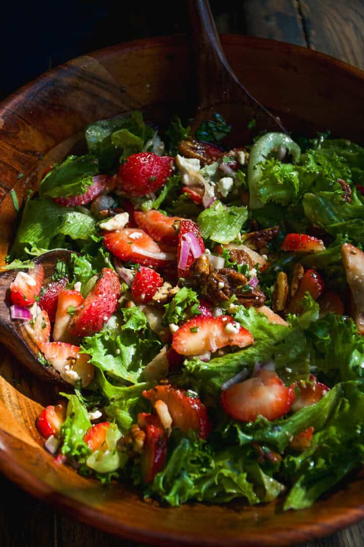 A wooden bowl filled with a fresh salad of leafy greens, sliced strawberries, pecans, red onions, and crumbled cheese, mixed with a wooden spoon.