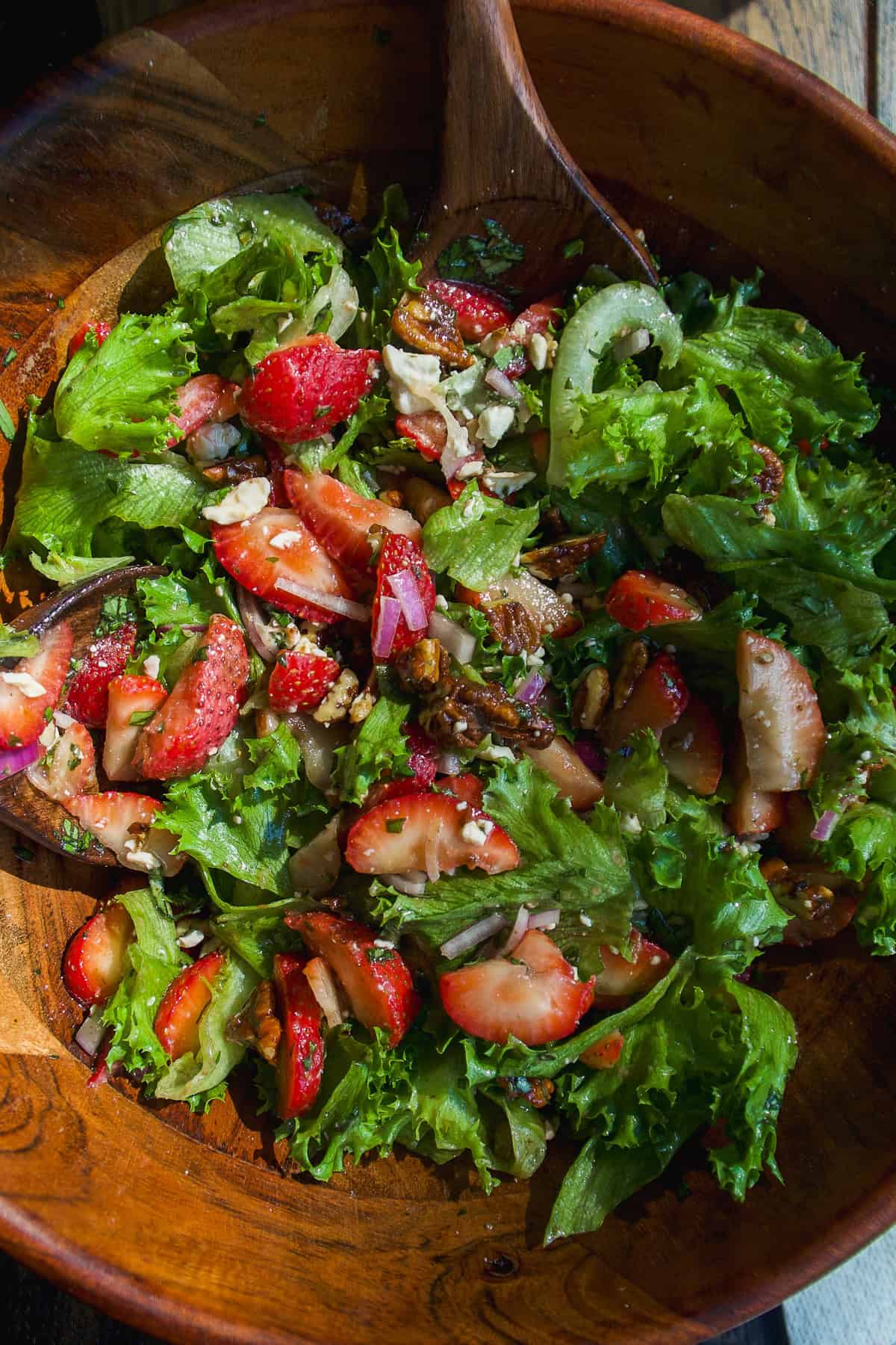 A wooden bowl filled with a fresh salad of mixed greens, sliced strawberries, red onions, nuts, and crumbled cheese, with a wooden spoon partially visible.