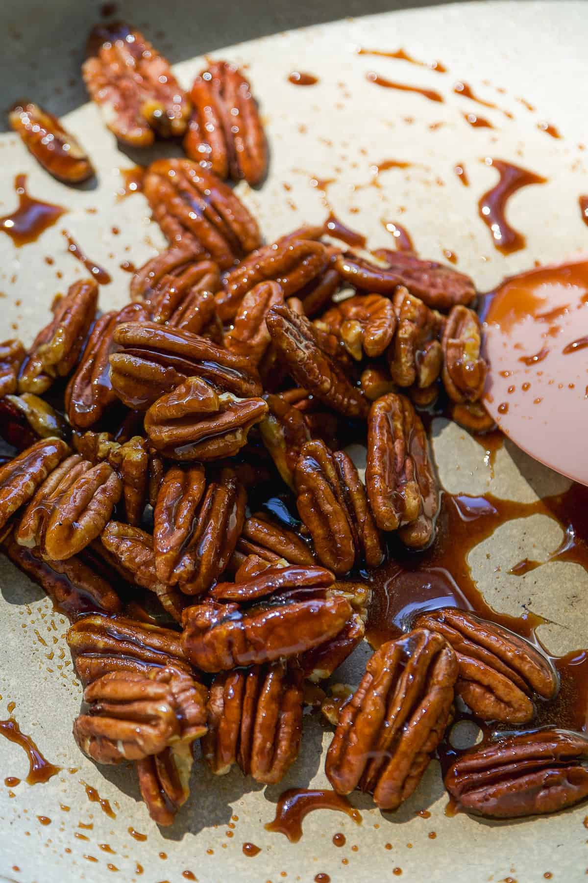 A close-up of glazed pecan halves in a pan, coated with a shiny, caramelized syrup. A rubber spatula is partially visible on the right side of the image.