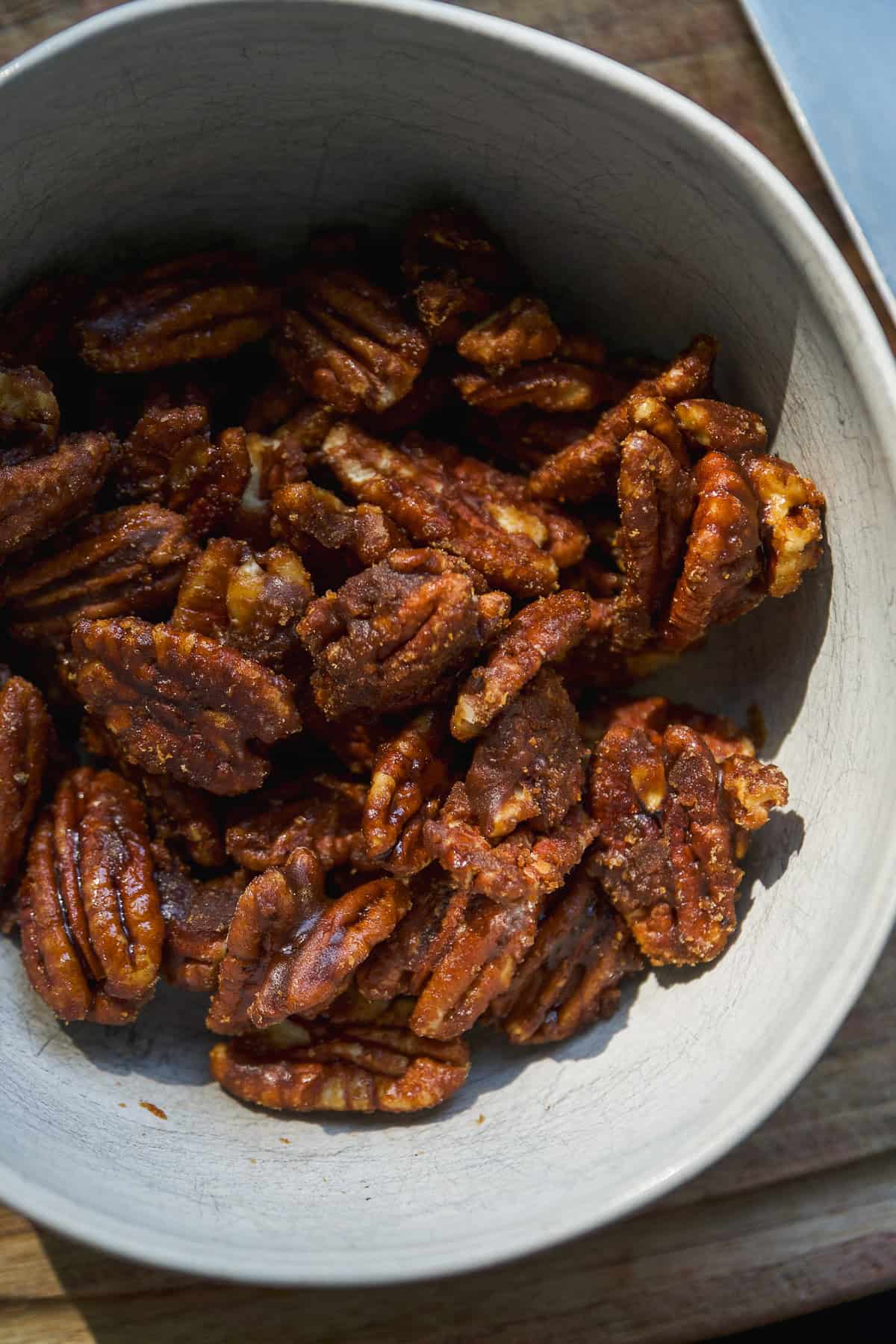 A close-up of a white bowl filled with candied pecans, showing their glossy, caramelized coating and textured surfaces, sitting on a wooden surface.