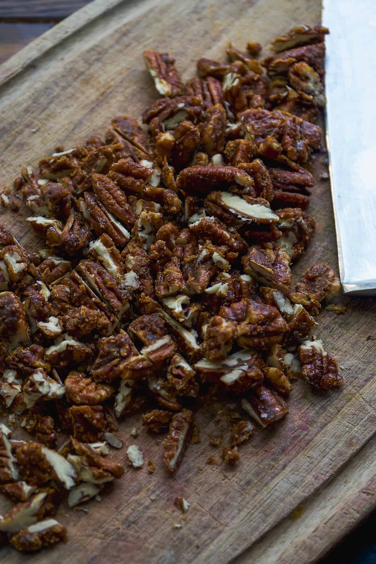 Chopped roasted pecans on a wooden cutting board with a large knife beside them.