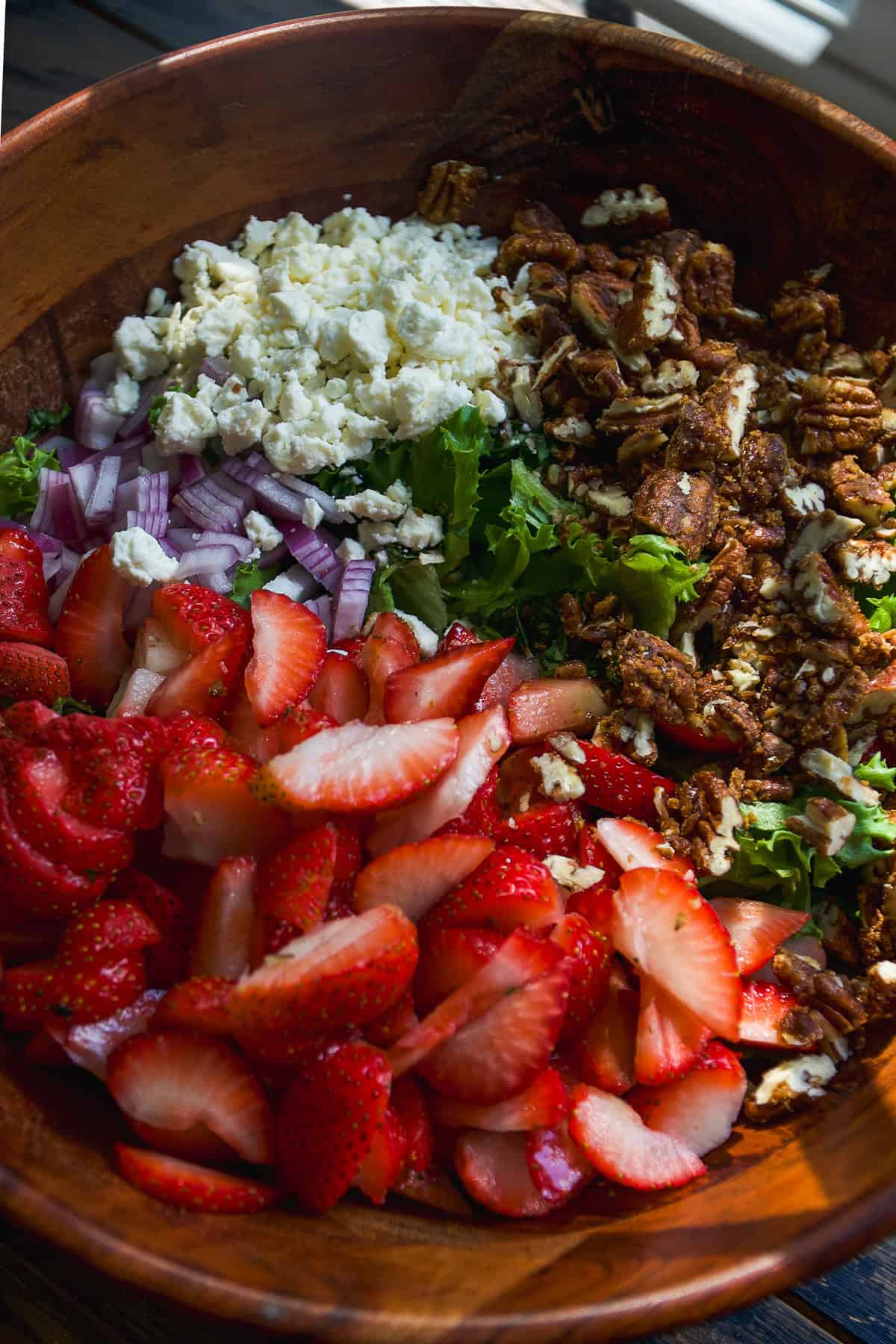 A wooden bowl filled with sliced strawberries, chopped red onions, crumbled feta cheese, leafy greens, and chopped pecans, ready to be mixed into a salad.