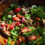 A wooden bowl filled with fresh salad featuring leafy greens, sliced strawberries, red onions, nuts, and chunks of cheese, tossed together with a wooden serving spoon.