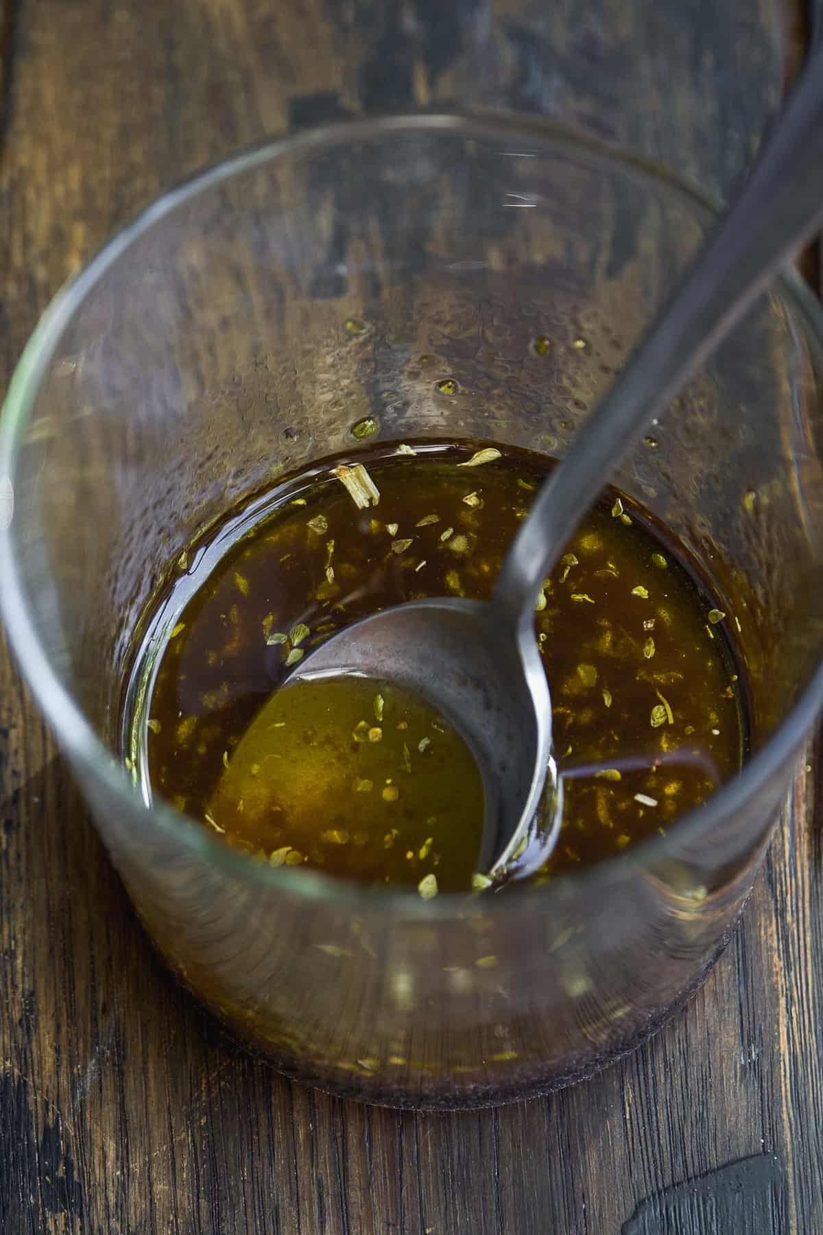 A glass cup filled with a brown, oily liquid containing herbs and seasonings, with a metal spoon resting inside. The cup sits on a rustic wooden surface.