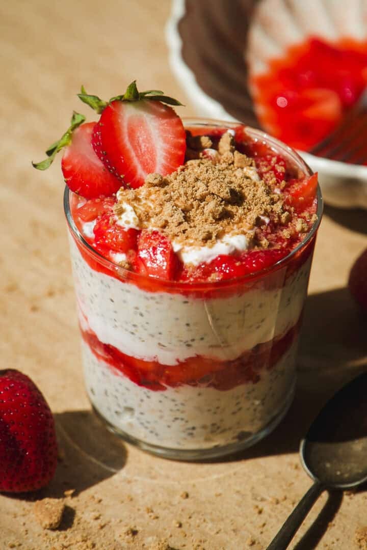 A glass filled with chia pudding layered with yogurt and fresh strawberries, topped with sliced strawberries and a sprinkle of crumbly topping, sits on a beige surface next to a spoon and whole strawberries.