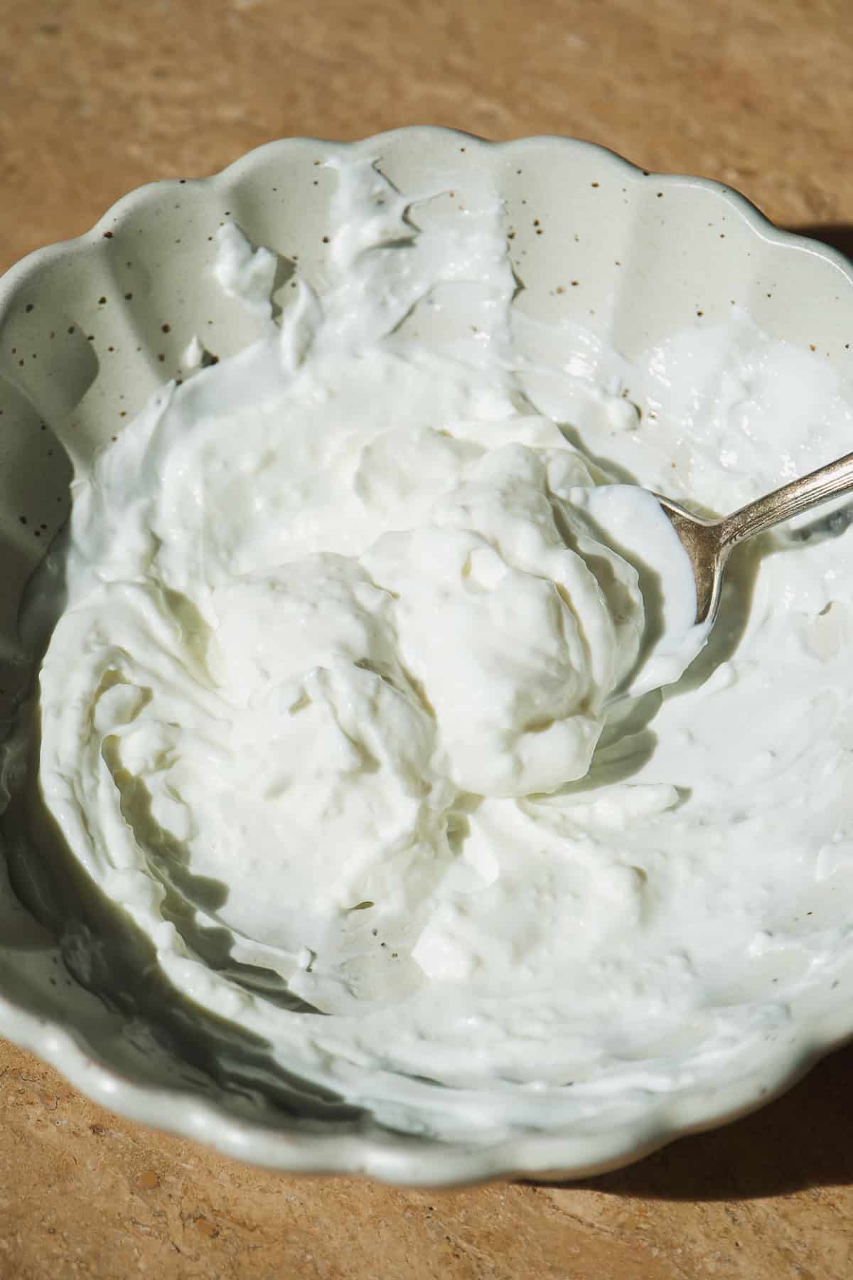A close-up of a bowl filled with thick, creamy white yogurt or sour cream, with a spoon resting inside. The bowl sits on a light brown surface.