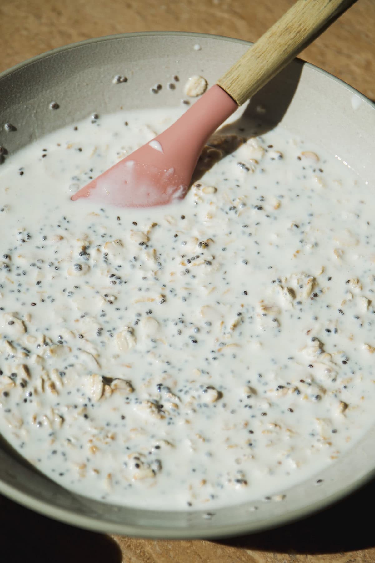 A bowl of overnight oats mixed with chia seeds and milk, stirred with a pink spatula on a wooden surface.