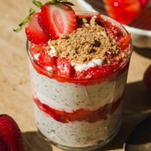 A glass filled with layered chia pudding, yogurt, sliced strawberries, and a crumb topping, garnished with halved strawberries. A whole strawberry and a spoon are beside the glass on a tan surface.