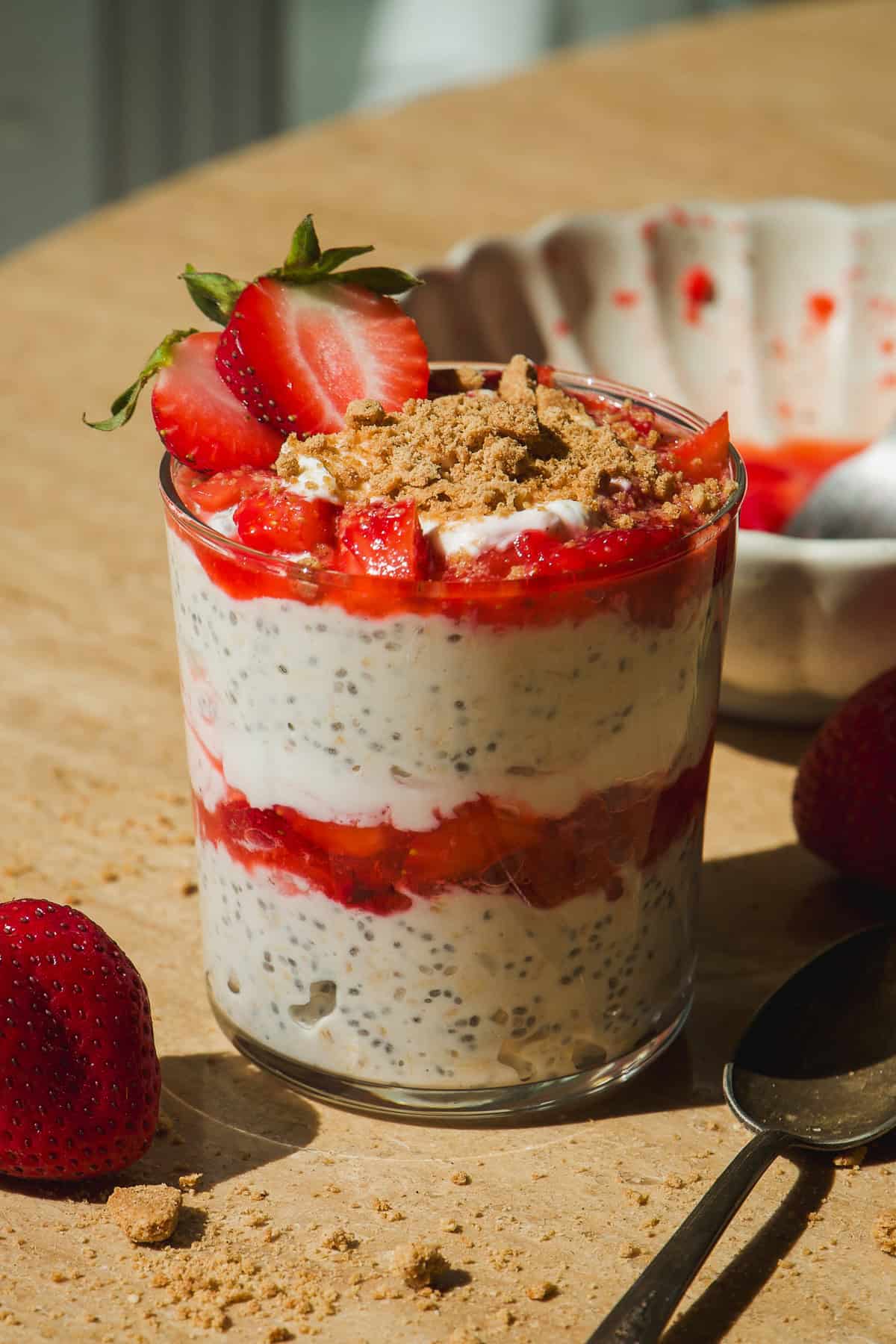 A glass filled with layered chia pudding, strawberries, and crumbled topping, garnished with fresh strawberries. A bowl with strawberry sauce and a spoon are beside the glass on a light surface.