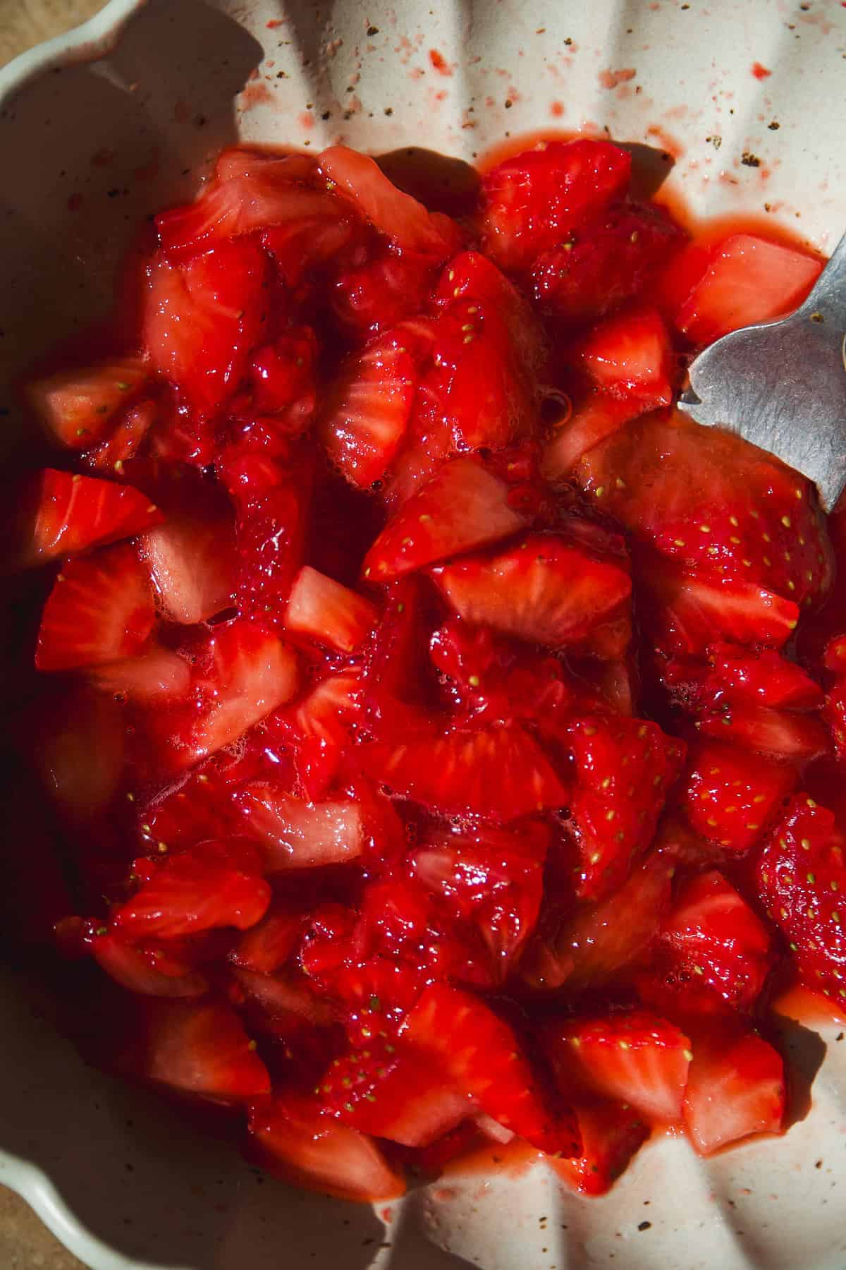 Close-up of freshly chopped strawberries in a bowl, mixed with their own juices. A spoon is partially visible at the edge of the bowl, suggesting the strawberries have been stirred or prepared for a dish.