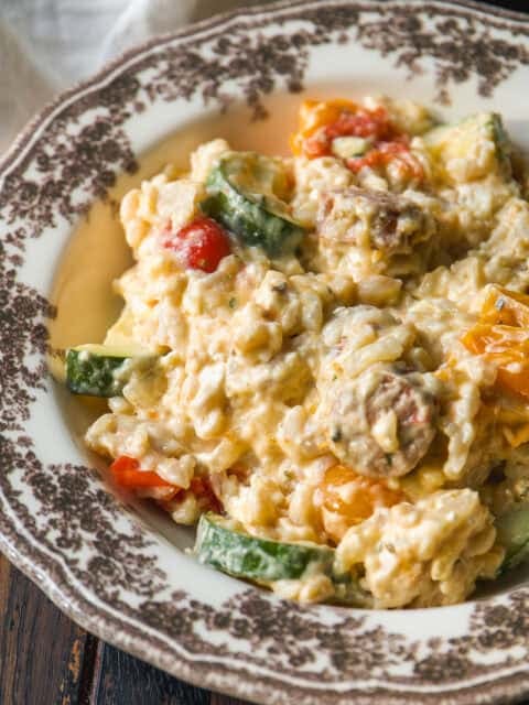 Creamy rice dish with zucchini, cherry tomatoes, and mushrooms, served on a vintage floral-patterned plate atop a rustic wooden table.